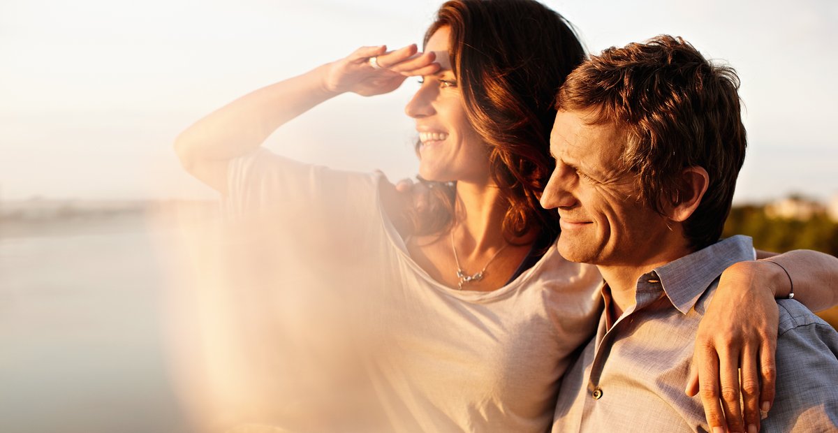 A couple looks from the sun deck of a river cruise ship at the Rhine in the sunshine. The woman shields her eyes with her hand to look into the distance.
