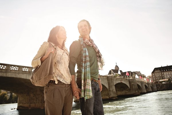 A couple is walking along the banks of the Rhine in Basel in the sunshine. In the background, there is a stone bridge that crosses the river.