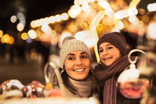 Woman and child with hat and scarf smiling at a Christmas market at night with string lights in the background.