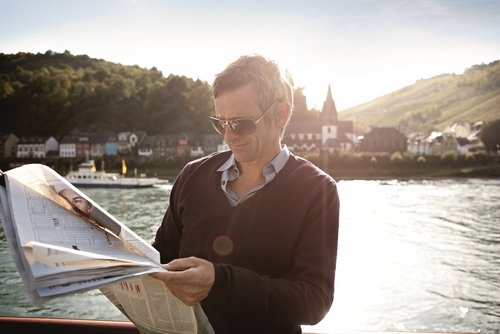 A man with sunglasses is reading a newspaper on board a river cruise ship in the sunshine. In the background is the Rhine with a city on the shore and a hilly landscape.