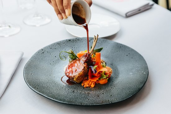 Sauce is poured over an artfully arranged rack of lamb with carrots and vegetables on a black plate.