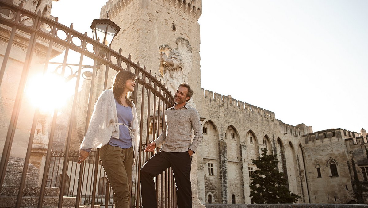 A couple stands in the old town of Avignon in front of a fence at a historic building. In the background, the sun is setting.