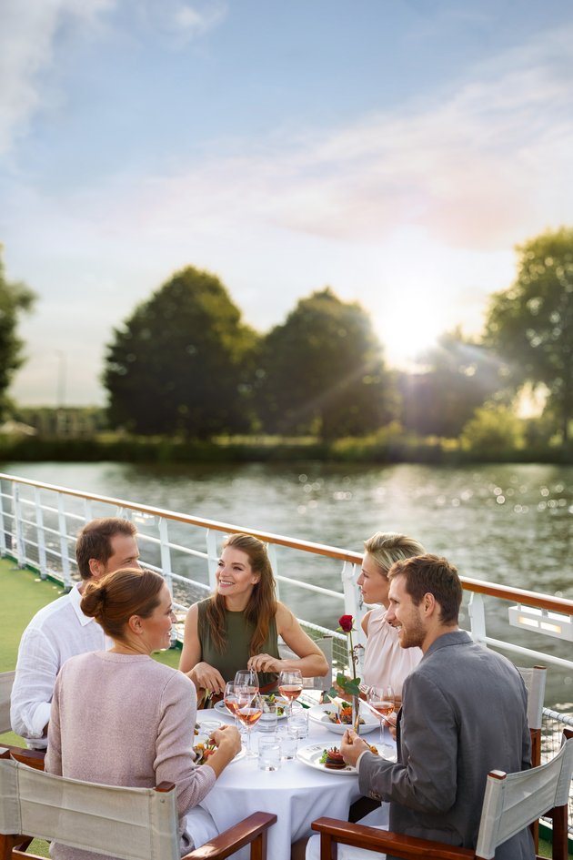 Yvonne Catterfeld and four other people are sitting at a set table on a sun deck by the water, enjoying a meal at sunset.