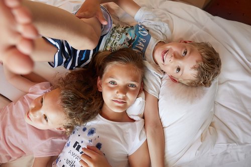 Three children lie close together on a bed, two girls and a boy, smiling and relaxed.