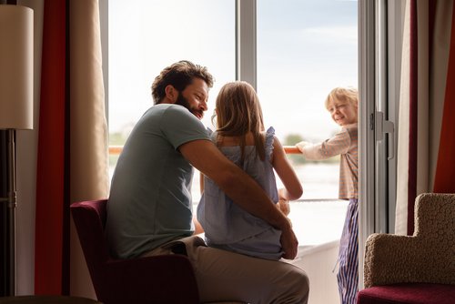 A father is sitting with his child in a family cabin on a river cruise ship in front of the balcony. A second child is standing on the balcony.