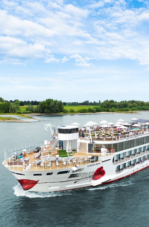 The A-ROSA SENA sails along the Rhine, the area with deck chairs and sunshades is well attended, with the riverbank featuring green landscapes and trees in the background.