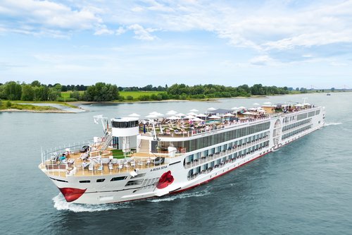 The A-ROSA SENA sails along the Rhine, the area with deck chairs and sunshades is well attended, with the riverbank featuring green landscapes and trees in the background.