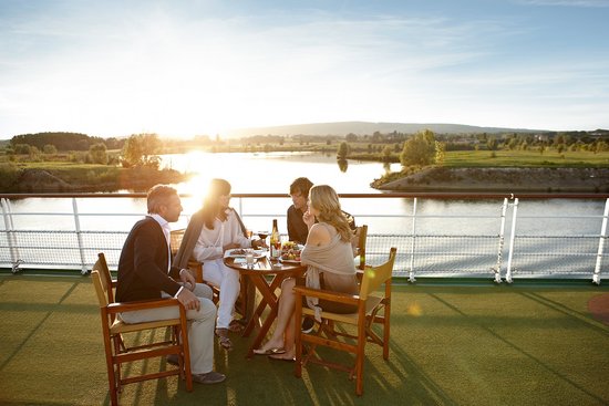 Two couples sit at a table on the sun deck, enjoying wine and something to eat at sunset, surrounded by green riverbank landscapes and a warm evening atmosphere.
