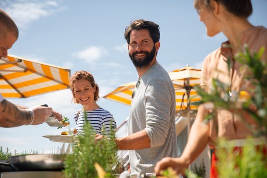 Group of people at an outdoor BBQ, a woman holding a plate, a man smiling at the camera, yellow and white sun umbrellas in the background.