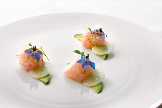 Three small portions of raw fish with thin cucumber slices and blue edible flowers arranged on a white plate.