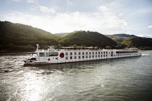 A ship from the A-ROSA Rhine fleet glides along the Rhine in the afternoon, with green, wooded hills and small villages in the background on the shore.