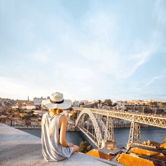 A woman with a white hat and a grey dress sits on a wall and looks at the Dom Luís I steel arch bridge over the river in Porto.