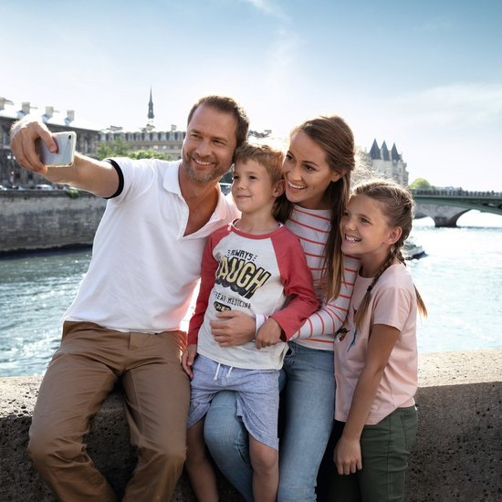 Family with father, mother, son, and daughter takes a selfie on a bridge over the river in Paris in sunny weather.