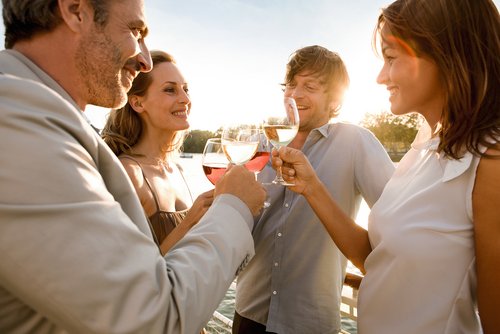 Two couples stand at the railing of a riverboat at sunset, clinking glasses of wine. In the background, green trees on the shore and the river.