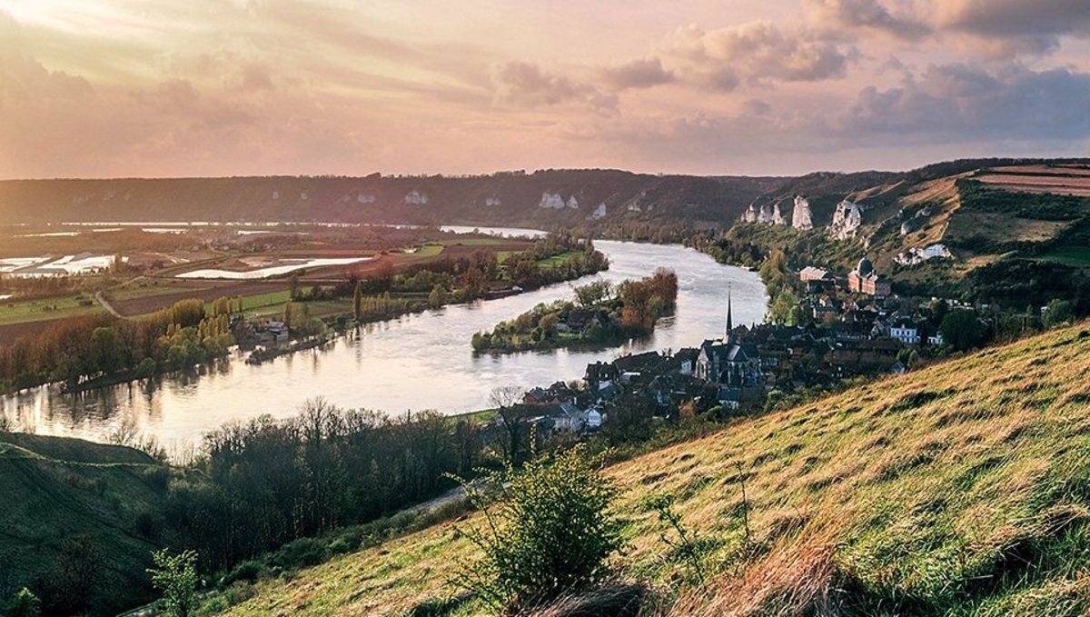 River Seine, winding through green hills and the village of Les Andelys with its church and rocky landscape at sunset.