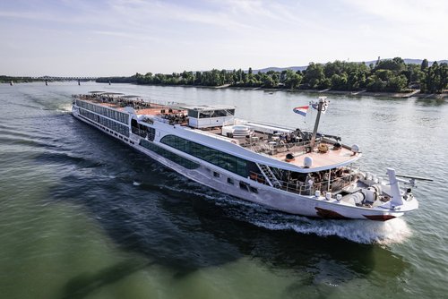 A river cruise ship sails along a wooded bank on the Rhine. The sun deck is equipped with a whirlpool, sun loungers, and sun sails.